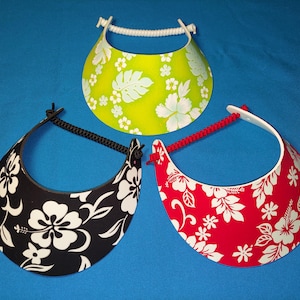 May include: Three sun visors featuring a floral pattern. One visor is lime green with white flowers, one is black with white flowers, and one is red with white flowers. Each visor has a curved brim and an adjustable cord strap at the back.