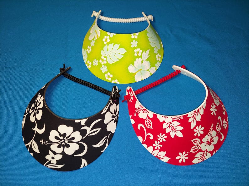 May include: Three sun visors featuring a floral pattern. One visor is lime green with white flowers, one is black with white flowers, and one is red with white flowers. Each visor has a curved brim and an adjustable cord strap at the back.