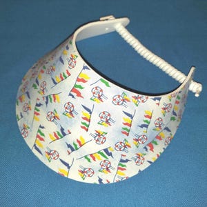 May include: A white plastic sun visor featuring a repeating pattern of colorful nautical flags and life preservers. The visor has a curved brim and an adjustable white coiled plastic strap at the back to secure the headpiece in place.