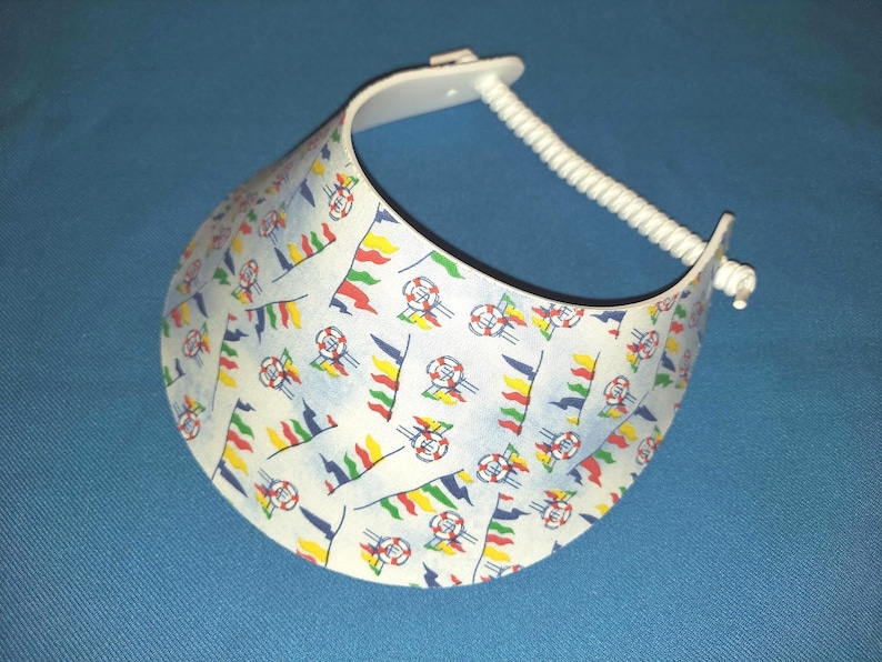 May include: A white plastic sun visor featuring a repeating pattern of colorful nautical flags and life preservers. The visor has a curved brim and an adjustable white coiled plastic strap at the back to secure the headpiece in place.