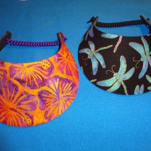 May include: Two sun visors with curved brims and adjustable back straps. One visor features an orange and purple hibiscus floral pattern with a purple strap, while the other has a black background with a blue and green dragonfly print and a black strap.