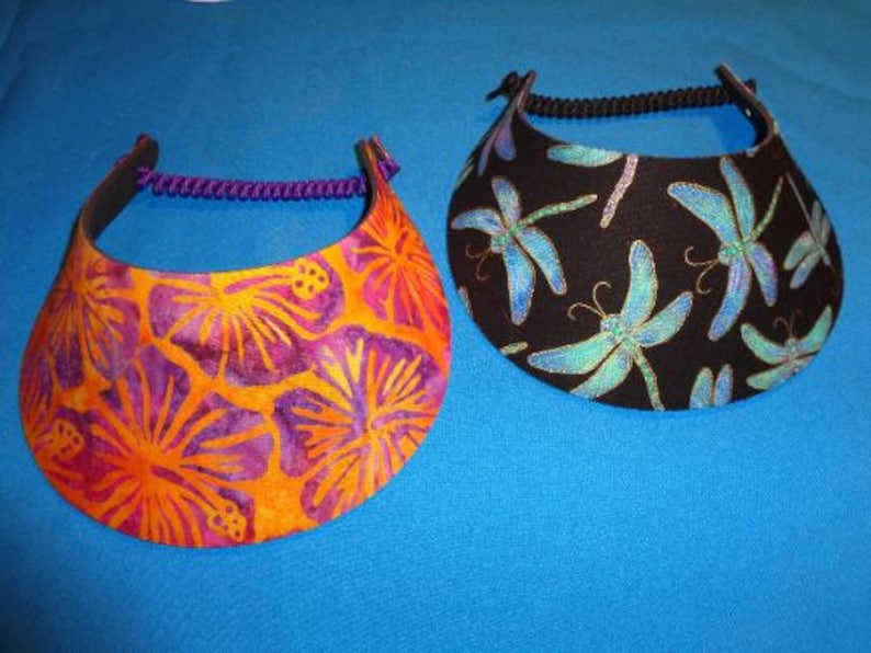 May include: Two sun visors with curved brims and adjustable back straps. One visor features an orange and purple hibiscus floral pattern with a purple strap, while the other has a black background with a blue and green dragonfly print and a black strap.