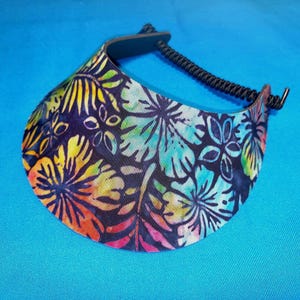 May include: A sun visor featuring a vibrant, multicolored floral pattern with shades of yellow, orange, pink, teal, and blue against a black background. The visor includes a black, adjustable, braided cord strap for securing the headpiece in place.