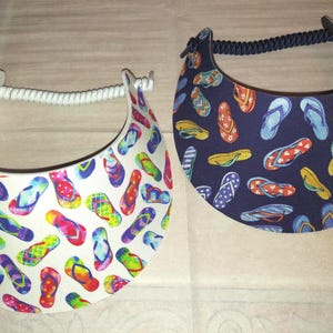 May include: Two sun visors featuring a repeating flip-flop pattern. One visor has a white background with multicolored flip-flops, while the other has a dark navy blue background with various flip-flop designs. Both include a coiled elastic strap.