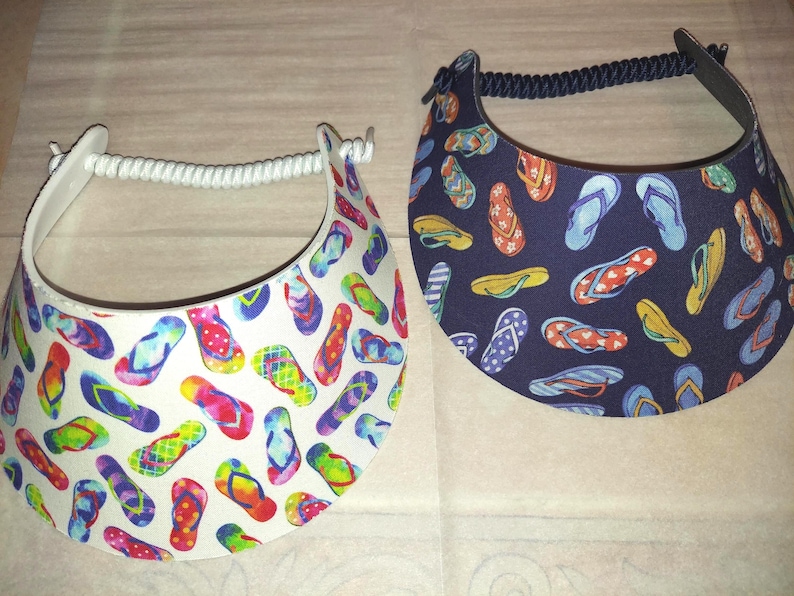 May include: Two sun visors featuring a repeating flip-flop pattern. One visor has a white background with multicolored flip-flops, while the other has a dark navy blue background with various flip-flop designs. Both include a coiled elastic strap.
