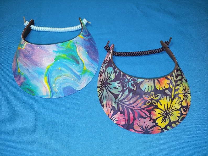 May include: Two sun visors with curved brims. The left visor features a blue, purple, and yellow marbled paint pattern with a light blue cord. The right visor displays a dark floral print with yellow, pink, and teal flowers and a black cord.