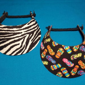 May include: Two sun visors with black coiled elastic headbands. One visor features a black and white zebra stripe pattern, while the other visor has a black background decorated with a colorful pattern of various flip-flop sandals.