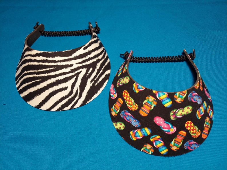 May include: Two sun visors with black coiled elastic headbands. One visor features a black and white zebra stripe pattern, while the other visor has a black background decorated with a colorful pattern of various flip-flop sandals.