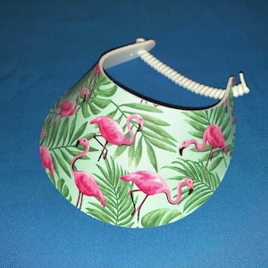 May include: A sun visor featuring a light green background with a repeating pattern of bright pink flamingos and dark green tropical palm leaves. The visor includes a white plastic band with a coiled, adjustable strap for securing the headpiece.