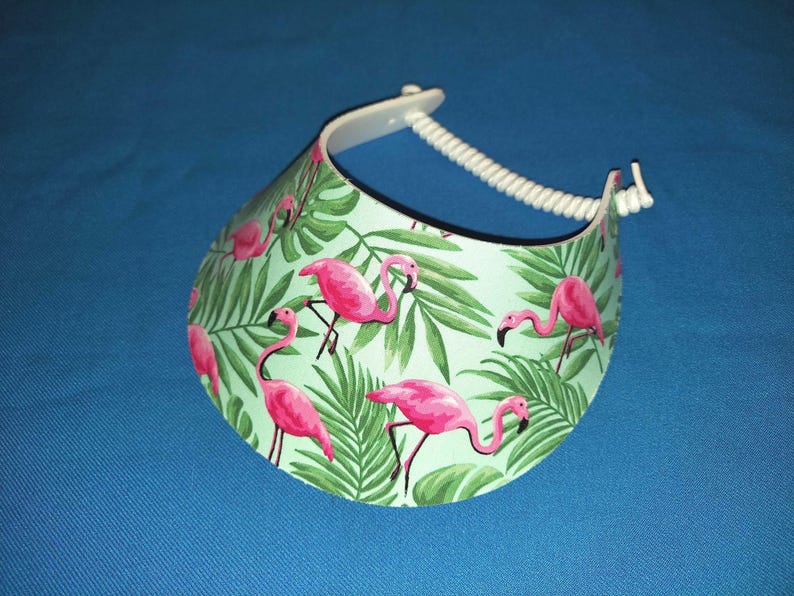 May include: A sun visor featuring a light green background with a repeating pattern of bright pink flamingos and dark green tropical palm leaves. The visor includes a white plastic band with a coiled, adjustable strap for securing the headpiece.