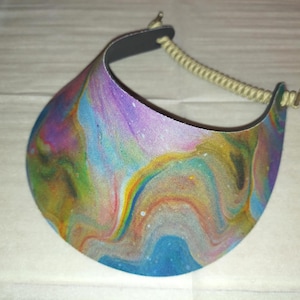May include: A sun visor featuring a vibrant, multi-colored fluid acrylic paint pour design with swirling patterns of blue, purple, yellow, and orange. The visor includes a beige coiled cord strap attached to the sides with small metal hardware.