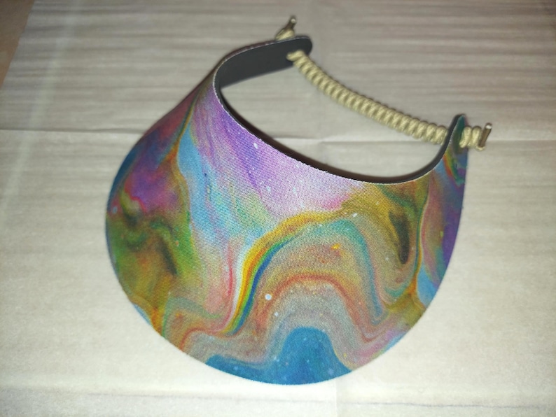 May include: A sun visor featuring a vibrant, multi-colored fluid acrylic paint pour design with swirling patterns of blue, purple, yellow, and orange. The visor includes a beige coiled cord strap attached to the sides with small metal hardware.