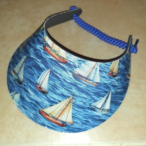 May include: A sun visor featuring a repeating pattern of sailboats on a blue ocean background. The visor has a curved brim and a blue coiled cord strap that connects the two sides, providing an adjustable fit for the wearer's head.