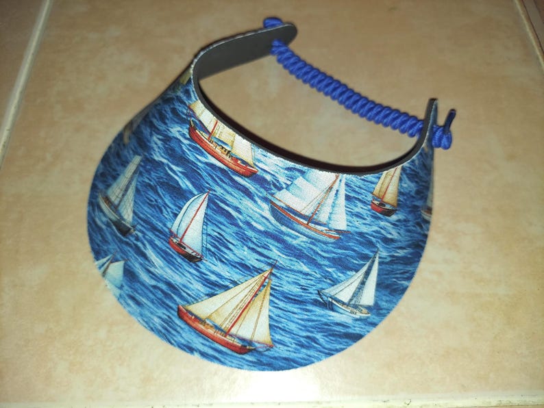 May include: A sun visor featuring a repeating pattern of sailboats on a blue ocean background. The visor has a curved brim and a blue coiled cord strap that connects the two sides, providing an adjustable fit for the wearer's head.