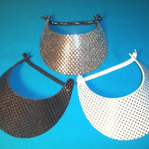 May include: Three sun visors in silver, black, and white, each featuring a textured, perforated surface pattern and a flexible, coiled headband design. These headwear items are arranged in a triangular formation, showcasing their distinct colors.