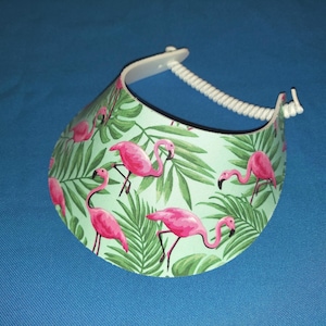 May include: A sun visor featuring a light green background with a repeating pattern of bright pink flamingos and dark green tropical palm leaves. The visor has a white plastic brim edge and a white coiled plastic strap for an adjustable fit.