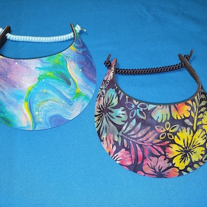 May include: Two sun visors with curved brims. The left visor features a blue, purple, and yellow marbled paint pattern with a light blue cord. The right visor displays a dark floral print with yellow, pink, and teal flowers and a black cord.