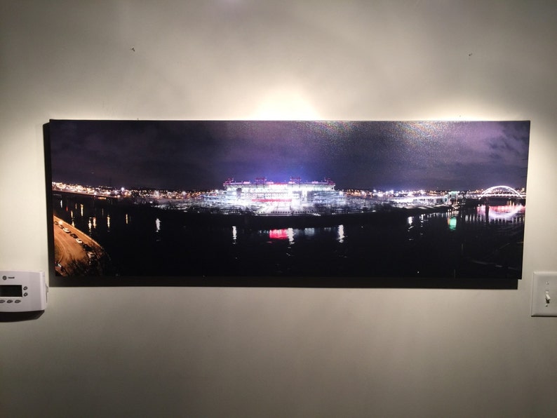 May include: A panoramic photograph of a city skyline at night. The city is lit up with lights, and a large stadium is visible in the center of the image. The stadium is surrounded by a body of water, and a bridge is visible in the distance.