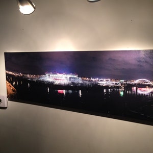 May include: A panoramic photograph of a city skyline at night, featuring a large stadium lit up with lights. The stadium is reflected in the water below, and there are several bridges in the distance.