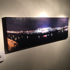 May include: A panoramic nighttime photograph of a city skyline with a large stadium lit up in the distance. The stadium is reflected in the water below. The city lights are visible in the background.