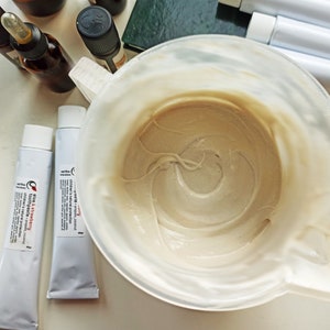May include: Two white tubes of toothpaste with red and black text. The tubes are labeled "Clay & Strawberry Toothpaste" and "Charcoal + Propolis Coconut Toothpaste". A white bowl with a beige paste inside.