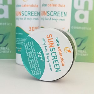 Puede incluir: Un pequeño tarro de crema solar de aloe vera y caléndula con una etiqueta blanca que dice "aloe calendula sunscreen daily face & body cream 30spf". El tarro está sobre una superficie blanca.