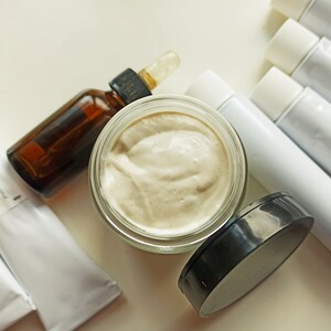 May include: A close-up of a glass jar filled with a light beige cream, a brown glass bottle with a dropper, and four white tubes with black caps. The cream is thick and smooth, and the bottle and tubes are all neatly arranged on a white surface.