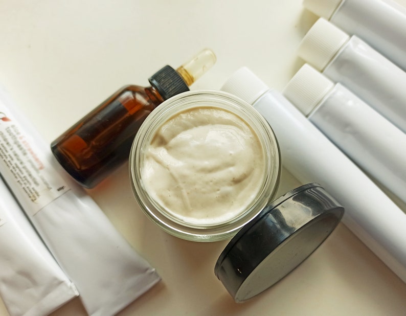 May include: A close-up of a glass jar filled with a light beige cream, a brown glass bottle with a dropper, and four white tubes with black caps. The cream is thick and smooth, and the bottle and tubes are all neatly arranged on a white surface.