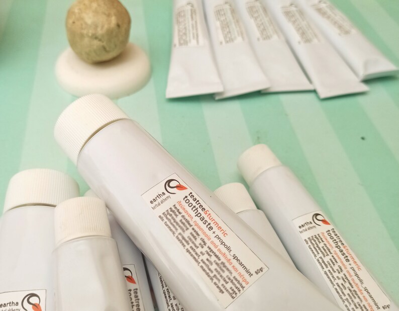 May include: Close-up of several white tubes of tea tree and turmeric toothpaste with propolis and spearmint. The tubes are labeled "eartha herbal alchemy" and have a list of ingredients and benefits.
