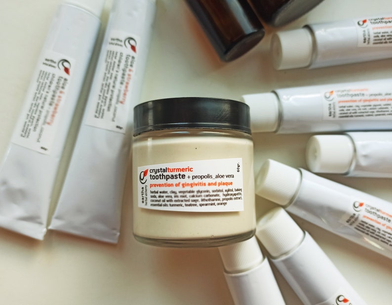 May include: A glass jar of crystal turmeric toothpaste with propolis and aloe vera. The jar is labeled with the text "crystalturmeric toothpaste + propolis aloe vera prevention of gingivitis and plaque".
