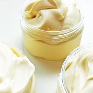 Op de afbeelding: Drie glazen potten gevuld met witte en gele crème. De crème is dik en romig, en de potten zijn van helder glas.
