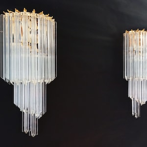 May include: Two clear glass wall sconces with a gold-coloured top and a tiered design. The sconces are composed of vertical glass rods, creating a chandelier-like appearance. The background is a solid black.