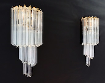 Peut inclure: Deux appliques murales en verre transparent avec un dessus doré et un design à plusieurs niveaux. Les appliques sont composées de tiges de verre verticales, créant un aspect de lustre. Le fond est noir uni.