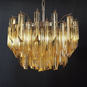 Pode incluir: Um candeeiro de cristal dourado com múltiplos níveis de prismas de vidro transparente. Os prismas são dispostos em cascata, criando um efeito cintilante.