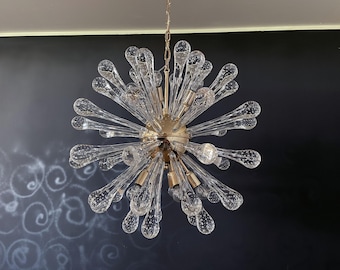 May include: A modern chandelier with a gold-colored central sphere and numerous clear, teardrop-shaped glass elements radiating outwards. The fixture is suspended from a chain and is designed to provide ambient lighting.