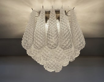 May include: A clear glass chandelier with a textured, teardrop-shaped design. The light fixture is suspended from a square metal frame and features multiple light sources. The glass has a bubble-like pattern, creating a unique lighting effect.