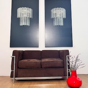 Może przedstawiać: Brązowa dwuosobowa sofa z chromowaną ramą i czerwony wazon z suszonymi kwiatami. Dwa pasujące szklane żyrandole są zamontowane na ciemnoniebieskich panelach na ścianie.