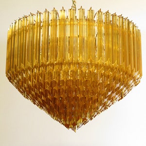 May include: A large, amber-coloured glass chandelier with a geometric design. The chandelier is made up of many long, thin pieces of glass that are arranged in a pattern of stars.
