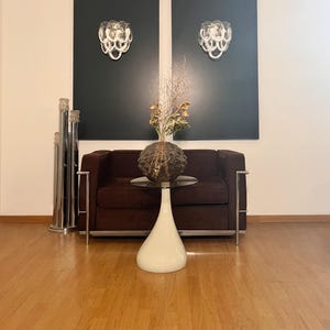 May include: Interior shot featuring a brown sofa, a white teardrop-shaped table, and a decorative vase with dried plants. Two dark gray panels on the wall each hold a decorative wall sconce. A tall, silver floor lamp stands to the left.
