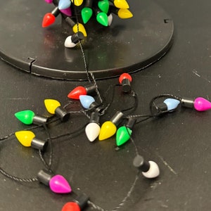 May include: A spool of colourful Christmas lights with a black cord. The lights are in a variety of colours, including red, green, blue, yellow, and white. The lights are arranged in a spiral on the spool.