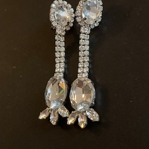 May include: A pair of silver dangle earrings with clear crystal stones. The earrings have a teardrop-shaped stone at the bottom and a smaller round stone at the top.