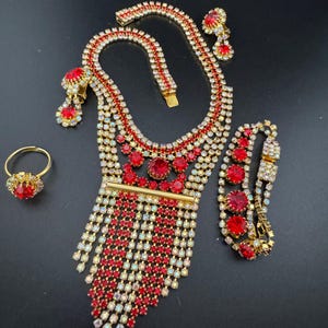 May include: A glamorous jewelry set featuring a necklace, bracelet, earrings, and ring. The set is gold-toned and adorned with red and clear rhinestones. The necklace has a cascading fringe design, and the ring has a large red stone.
