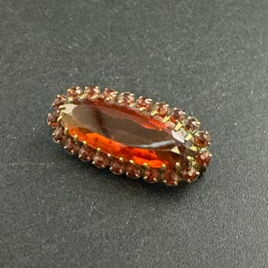 May include: An oval-shaped antique brooch with a large, faceted amber-colored center stone. The stone is surrounded by a border of smaller, reddish-brown gemstones set in a gold-toned metal frame. The brooch is a vintage piece.