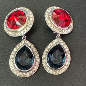Peut inclure: Une paire de boucles d'oreilles pendantes avec une pierre ronde rouge et une pierre en forme de larme bleue, toutes deux serties dans un métal argenté avec un halo de cristal transparent.
