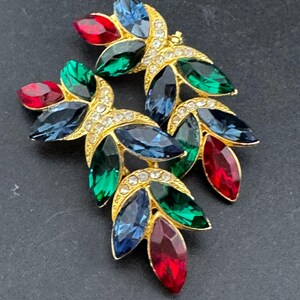 May include: A pair of gold-toned leaf-shaped brooches. Each brooch features a cluster of colorful, faceted gemstones in red, green, and blue. The gold setting is embellished with small, clear crystals, adding sparkle to the jewelry.