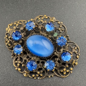 May include: A vintage brooch featuring a blue glass cabochon surrounded by smaller blue rhinestones in a gold tone filigree setting.