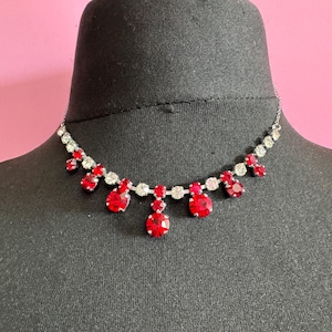 May include: A silver-toned necklace with red and clear gemstones. The necklace features a series of small, clear stones alternating with larger, teardrop-shaped red stones. The necklace is displayed on a black mannequin head against a pink background.
