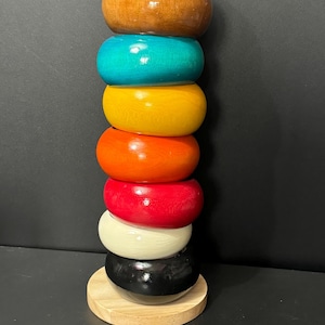 May include: A stack of eight wooden bangles in various colours, including brown, teal, yellow, orange, red, white, and black. The bangles are stacked on a wooden base.
