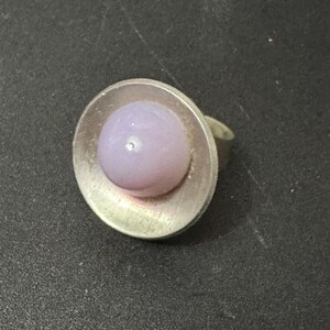 May include: A silver ring with a round, light purple glass stone set in the centre.