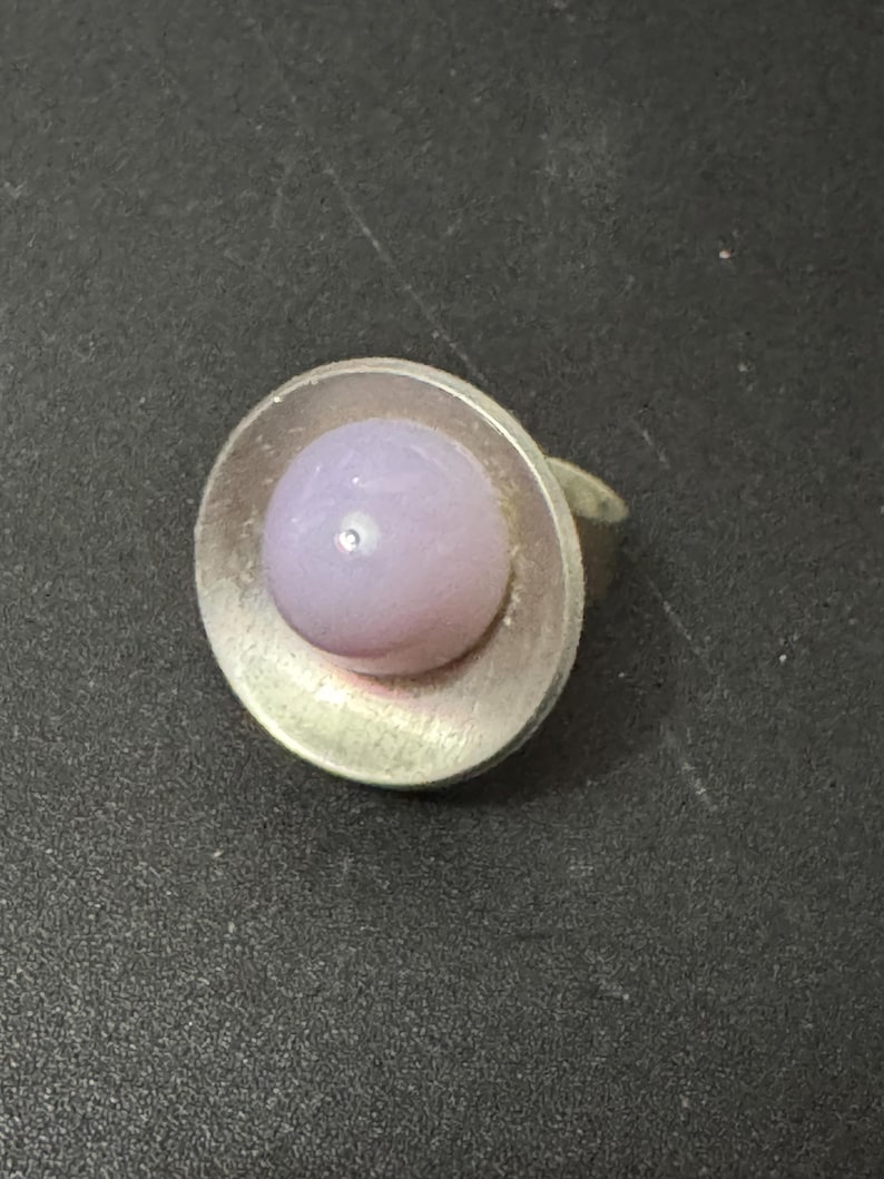 May include: A silver ring with a round, light purple glass stone set in the centre.